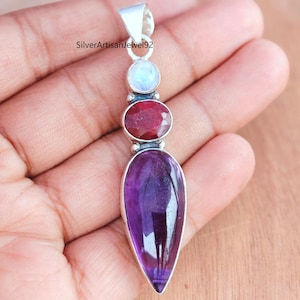 May include: A silver pendant with three gemstones: a round, pale blue moonstone, a round, deep red ruby, and a large, teardrop-shaped, deep purple amethyst. The pendant is held by a hand.