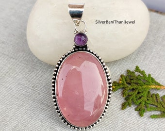Puede incluir: Un colgante de plata con una gran piedra ovalada de cuarzo rosa y una piedra redonda más pequeña de amatista. El colgante tiene un borde de cuentas y un aro para sujetar una cadena.