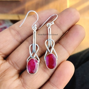 May include: A pair of silver dangle earrings with a red oval gemstone. The earrings feature a delicate knot design.