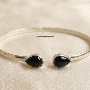 Puede incluir: Un brazalete plateado con dos piedras de ónix negro en forma de lágrima. El diseño abierto presenta piedras pulidas en cada extremo, sobre un fondo neutro. El texto "SilverArtisanJewel92" es visible.