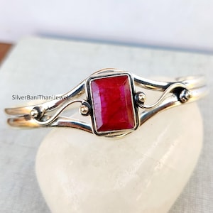 Può includere: Un braccialetto in argento con una pietra preziosa rossa incastonata in una cornice d'argento. Il braccialetto ha un design delicato e a spirale.