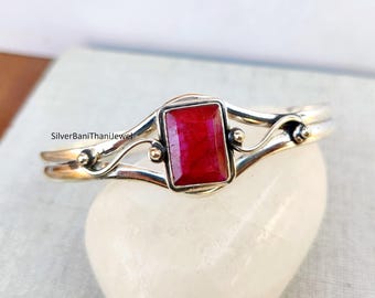 Puede incluir: Un brazalete de plata con una piedra preciosa roja engastada en un marco de plata. El brazalete tiene un diseño delicado y en espiral.