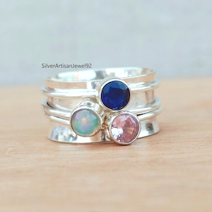 Può includere: Un anello d'argento con tre pietre preziose colorate: un blu intenso, un rosa chiaro e un'opale iridescente. L'anello ha un design a strati con finitura lucida.