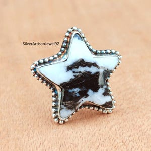 May include: A star-shaped ring with a white and black stone center. The ring has a silver beaded border and is set against a light brown surface. The text "SilverArtisanJewel92" is visible on the left side.