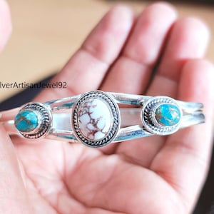 May include: A silver cuff bracelet with three oval turquoise and white stone settings. The central stone is white with brown marbling, flanked by two turquoise stones. The bracelet is held in a hand.