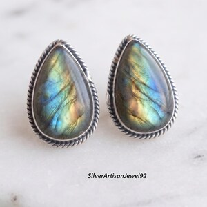 Puede incluir: Un par de pendientes de botón de labradorita en forma de lágrima engastados en plata de ley con un borde de cuerda trenzada. Cada piedra muestra destellos iridiscentes azules, dorados y verdes. El texto SilverArtisanJewel92 es visible abajo.