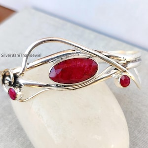 May include: A silver bangle bracelet with a delicate, interwoven design. The bracelet features three red gemstones, one large oval-shaped stone and two smaller round stones.
