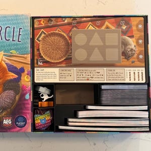 May include: A board game box titled "Knitting Circle: A Calico Game" featuring an orange cat sleeping in a basket of yarn. The box is open, revealing game components, including cards, game pieces, and a game board with a cat illustration.