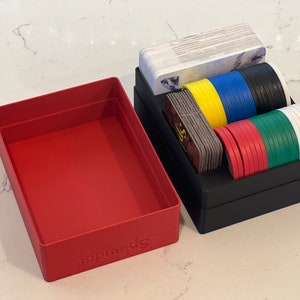May include: A red box with the word "Splendor" printed on the bottom, next to a black box with dividers for organizing game cards and tokens. The box contains a stack of cards, yellow, blue, black, red, green, and white tokens.