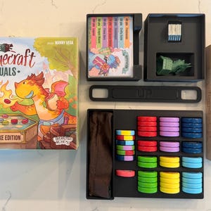 May include: Flamecraft Duels Deluxe Edition board game. The box features a dragon illustration. The game includes cards, tokens, and a dragon figurine. The tokens are in various colors, including red, blue, and green.
