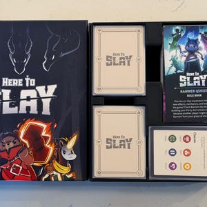 May include: An open board game box with the title "Here to Slay" on the box and cards. The box features illustrations of fantasy characters. Inside are card decks, a rule book, and other game components. The "Banner Quest" expansion is also visible.