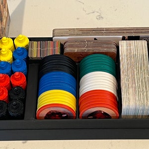May include: A black board game organizer tray containing stacks of colorful circular tokens in yellow, blue, red, and black, alongside two larger stacks of multi-colored circular chips, rectangular card decks, and small square tiles.