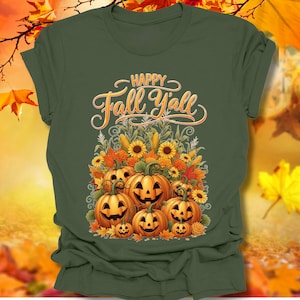 以下が含まれることがあります： 秋をテーマにしたグラフィックの緑色のTシャツ。デザインは、笑顔のカボチャ、ヒマワリ、装飾的なフォントの「Happy Fall Y'all」というテキストが特徴です。このシャツは半袖のクルーネックです。