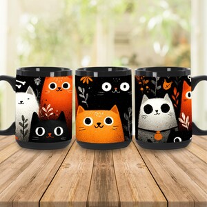 May include: Three black coffee mugs with cat illustrations. Each mug features a different design with cartoon cats in orange, white, and black, with floral accents. The mugs are on a wooden table.