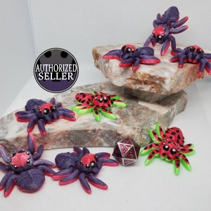 Puede incluir: Una colección de ocho figuritas de araña en miniatura pintadas a mano. Las arañas están pintadas en varios tonos de morado, rojo, negro y verde. Algunas arañas tienen manchas negras, mientras que otras tienen acentos verdes. Las arañas están dispuestas sobre una superficie blanca con una roca gris en el fondo.