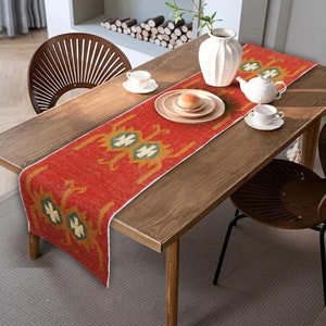 Op de afbeelding: Een rode tafelrunner met een geometrisch patroon in tinten goud en groen. De runner ligt op een houten tafel met twee stoelen en een vaas met bloemen.