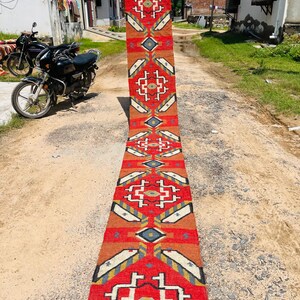 Puede incluir: Una alfombra larga y estrecha de color rojo, blanco y azul con un patr&oacute;n geom&eacute;trico. La alfombra est&aacute; tendida en un camino de tierra.