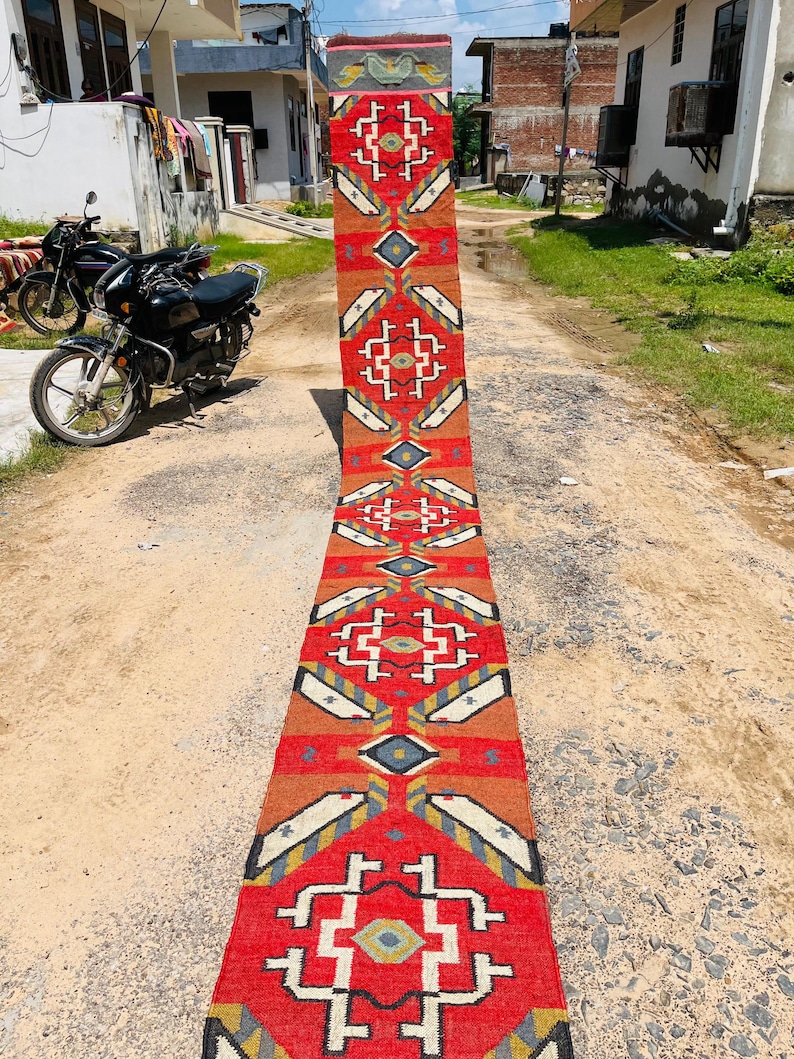 Puede incluir: Una alfombra larga y estrecha de color rojo, blanco y azul con un patr&oacute;n geom&eacute;trico. La alfombra est&aacute; tendida en un camino de tierra.