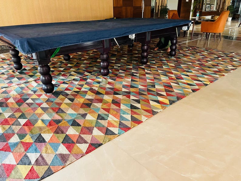 May include: A dark blue pool table cover with a wooden frame and legs. The table is set up on a colorful geometric patterned rug.