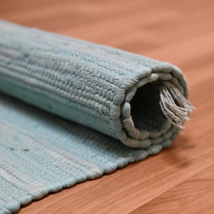 Op de afbeelding: Opgelrolde, lichtblauwe geweven vloerkleed met een getextureerd oppervlak en een gefranjerde rand. Het kleed is gedeeltelijk opgerold, waardoor de binnenkant en het geweven patroon zichtbaar zijn. De achtergrond is een houten oppervlak.