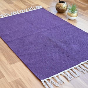 May include: A rectangular purple rug with white fringe edges. The rug is made of woven material and is placed on a wooden floor. Decorative items are in the background.