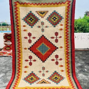 Pode incluir: Um tapete retangular com fundo creme e um padr&atilde;o geom&eacute;trico em tons de vermelho, amarelo, verde e azul. O tapete tem uma borda vermelha com franjas.