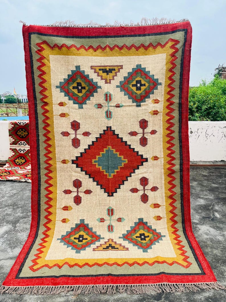 Pode incluir: Um tapete retangular com fundo creme e um padr&atilde;o geom&eacute;trico em tons de vermelho, amarelo, verde e azul. O tapete tem uma borda vermelha com franjas.