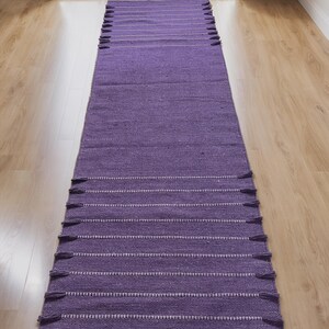 May include: A long, rectangular purple rug with white horizontal stripes and tassels on each end. The rug is made of a woven material and is placed on a light-colored wooden floor. The rug is a home decor item.
