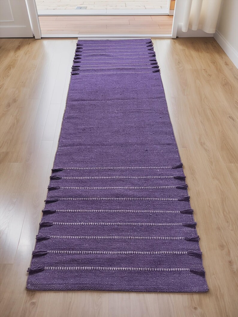 May include: A long, rectangular purple rug with white horizontal stripes and tassels on each end. The rug is made of a woven material and is placed on a light-colored wooden floor. The rug is a home decor item.