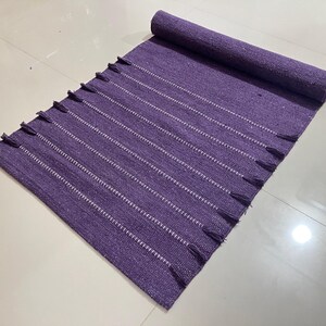 May include: A rolled-up, rectangular purple rug with white stripes and tassels on the ends. The rug is made of woven material and is placed on a light-colored tiled surface. The rug is a home decor item.