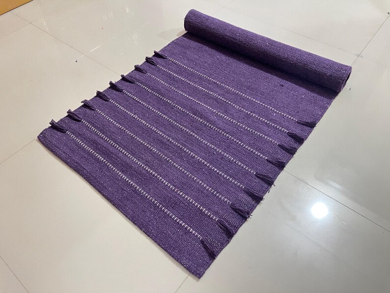 May include: A rolled-up, rectangular purple rug with white stripes and tassels on the ends. The rug is made of woven material and is placed on a light-colored tiled surface. The rug is a home decor item.