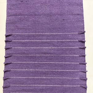 May include: A rectangular, woven purple rug with white horizontal stripes and fringe detailing. The rug is made of a textured material and is suitable for use in a variety of spaces. The rug's design is simple and elegant.