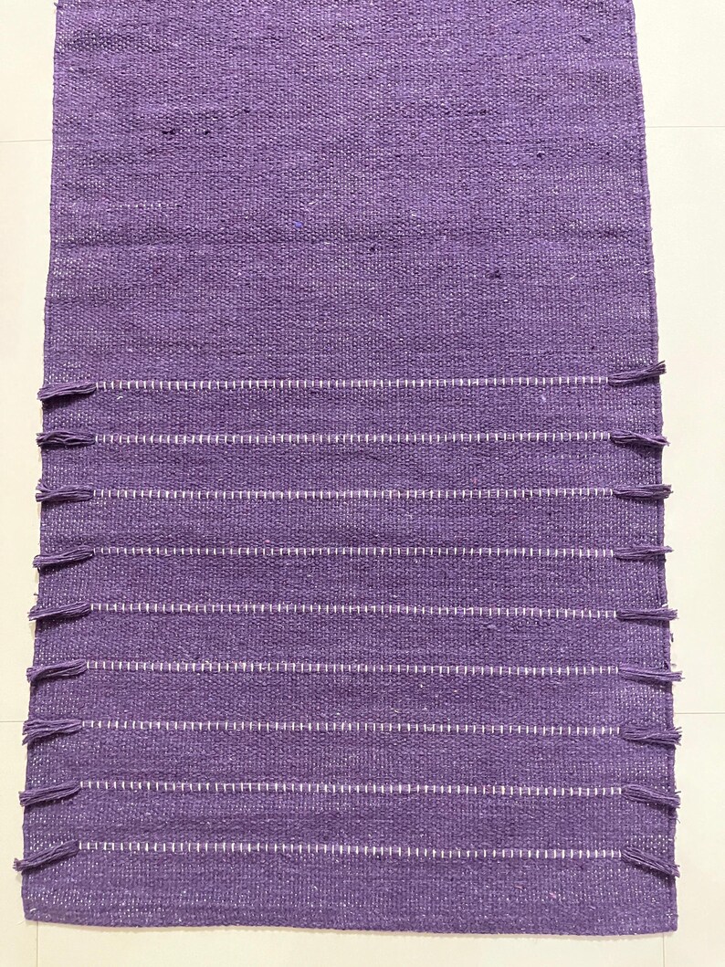 May include: A rectangular, woven purple rug with white horizontal stripes and fringe detailing. The rug is made of a textured material and is suitable for use in a variety of spaces. The rug's design is simple and elegant.
