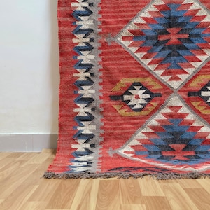 K&ouml;nnte beinhalten: Ein roter und blauer Teppich mit geometrischem Muster und Fransen. Der Teppich weist Rauten- und Zickzack-Designs in Rot-, Blau-, Wei&szlig;- und Graut&ouml;nen auf. Der Teppich liegt auf einem Holzboden.