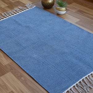 May include: A rectangular blue rug with fringed edges. The rug is made of woven material and has a textured appearance. The rug is placed on a wooden floor. Decorative items are visible in the background.