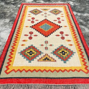 Pode incluir: Um tapete de cor creme com um padr&atilde;o geom&eacute;trico em tons de vermelho, amarelo, azul e verde. O tapete tem uma borda de franjas e &eacute; feito de fibras naturais.