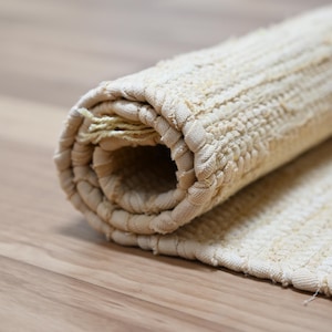 Puede incluir: Alfombra beige enrollada con un diseño tejido y texturizado. La alfombra está hecha de un material de color claro y está parcialmente desenrollada, revelando su construcción. La alfombra está sobre una superficie de madera.