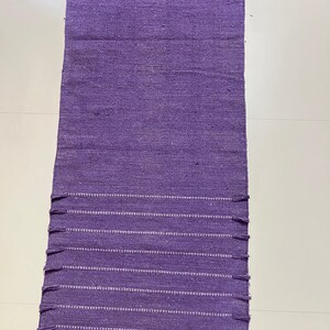 May include: A rectangular, woven purple rug with white horizontal stripes and fringe detailing on the ends. The rug is made of a textured material and is displayed on a white surface. The rug is a home decor item.