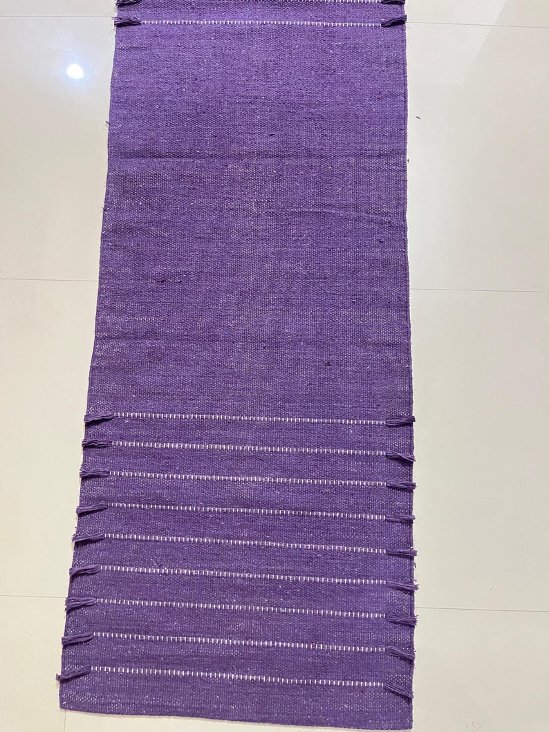 May include: A rectangular, woven purple rug with white horizontal stripes and fringe detailing on the ends. The rug is made of a textured material and is displayed on a white surface. The rug is a home decor item.