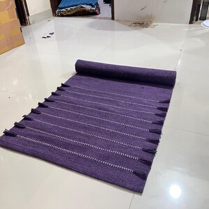 May include: A rolled-up, rectangular purple rug with white horizontal stripes and tassels along the edge. The rug is made of woven fabric and is partially unrolled on a white tiled floor.