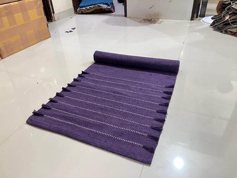 May include: A rolled-up, rectangular purple rug with white horizontal stripes and tassels along the edge. The rug is made of woven fabric and is partially unrolled on a white tiled floor.