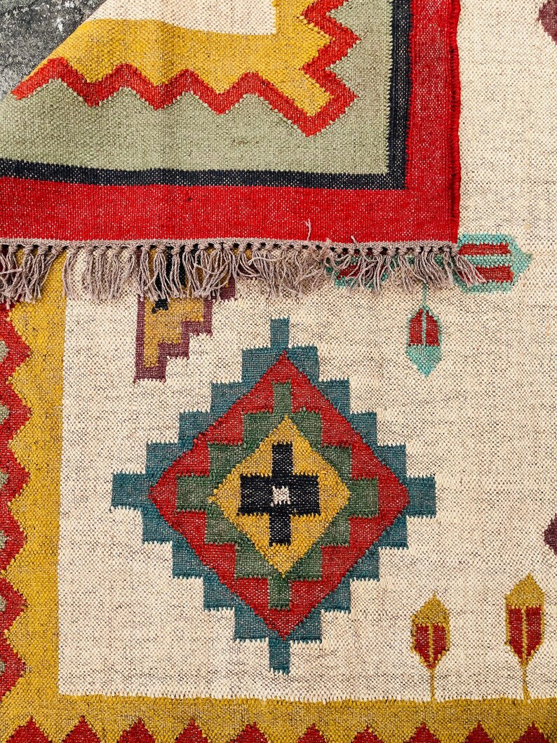 Pode incluir: Um tapete branco com um padr&atilde;o geom&eacute;trico colorido. O tapete tem uma borda de franjas e apresenta uma grande forma de diamante no centro com cores vermelho, verde, azul e amarelo.