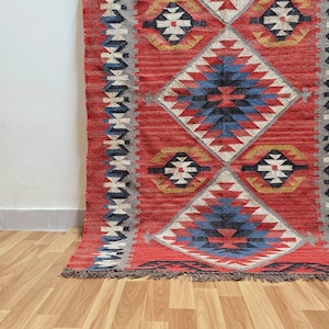 K&ouml;nnte beinhalten: Ein roter Teppich mit geometrischem Muster, mit Rauten- und Blumenmotiven. Der Teppich hat ein zentrales Rautenmuster in Blau, Wei&szlig; und Schwarz, umgeben von roten und grauen Bord&uuml;ren. Er hat einen Fransenrand und liegt auf einem Holzboden.