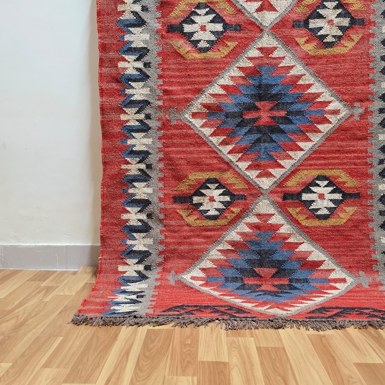 K&ouml;nnte beinhalten: Ein roter Teppich mit geometrischem Muster, mit Rauten- und Blumenmotiven. Der Teppich hat ein zentrales Rautenmuster in Blau, Wei&szlig; und Schwarz, umgeben von roten und grauen Bord&uuml;ren. Er hat einen Fransenrand und liegt auf einem Holzboden.