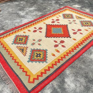 Pode incluir: Um tapete colorido com um padr&atilde;o geom&eacute;trico em tons de vermelho, amarelo, verde, azul e preto. O tapete tem uma borda de franjas e &eacute; feito de materiais tecidos.