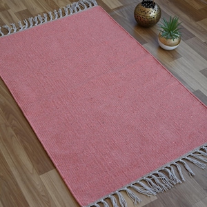 May include: A rectangular coral-colored rug with a woven texture and fringed edges. The rug is displayed on a wooden floor. Decorative items, including a gold-colored lantern and a small potted plant, are placed nearby.