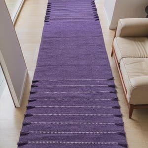 May include: A long, rectangular purple rug with white horizontal stripes and tassels on each end. The rug is made of a woven material and is placed on a light-colored wooden floor. The rug is in a hallway.