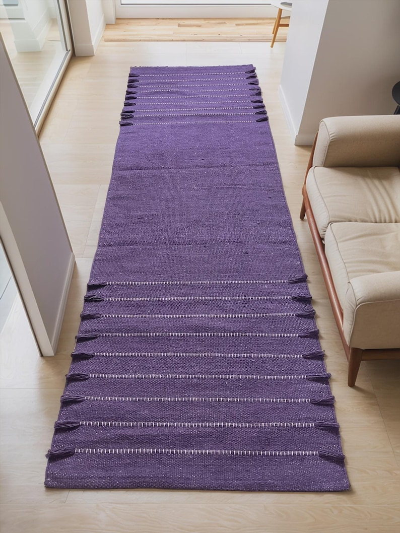 May include: A long, rectangular purple rug with white horizontal stripes and tassels on each end. The rug is made of a woven material and is placed on a light-colored wooden floor. The rug is in a hallway.