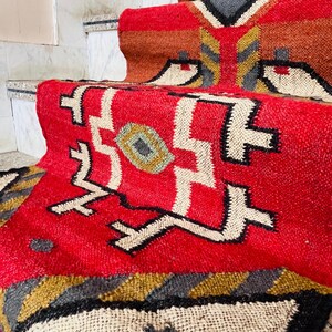 Puede incluir: Alfombra roja, blanca y negra con dise&ntilde;os geom&eacute;tricos. La alfombra est&aacute; hecha de lana tejida y tiene un borde con flecos.