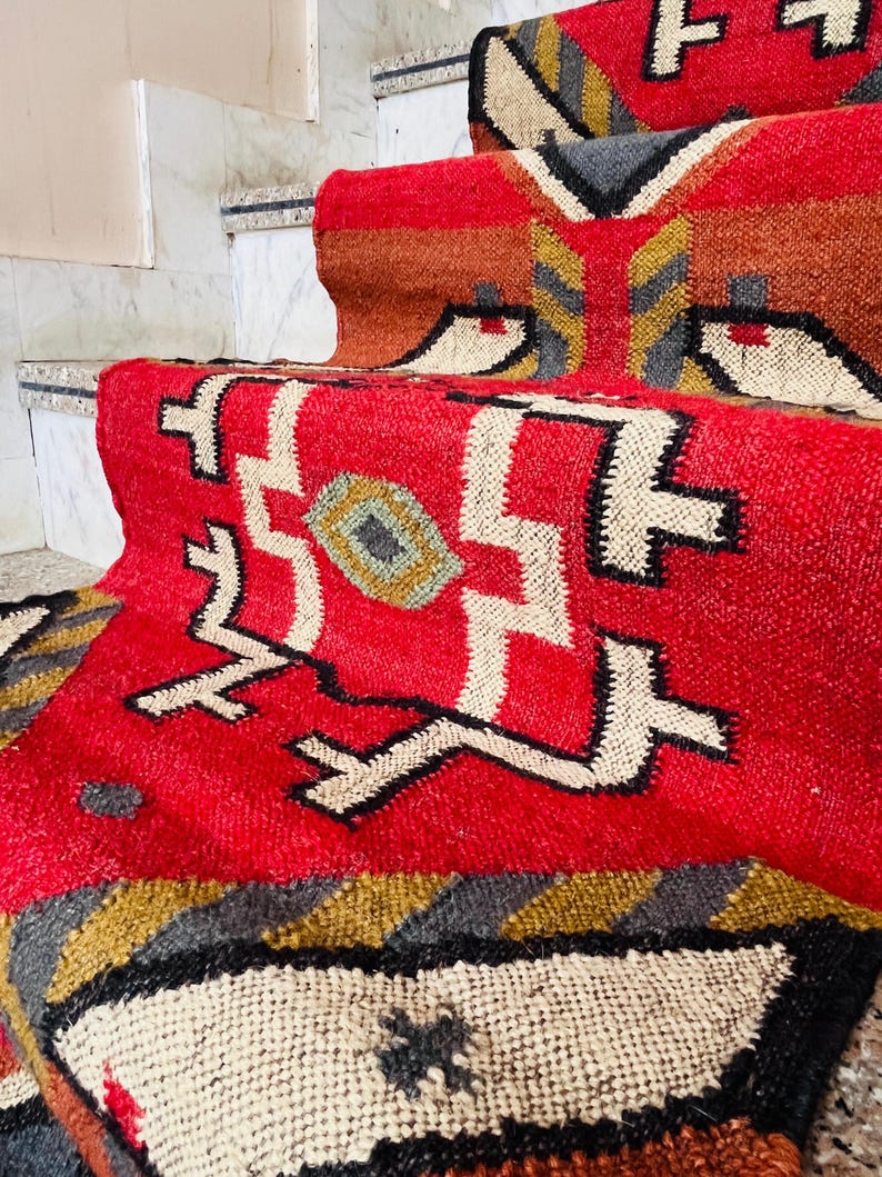 Puede incluir: Alfombra roja, blanca y negra con dise&ntilde;os geom&eacute;tricos. La alfombra est&aacute; hecha de lana tejida y tiene un borde con flecos.